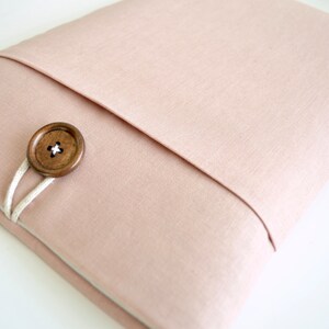 May include: A light pink fabric laptop sleeve with a wooden button closure and a white cord.