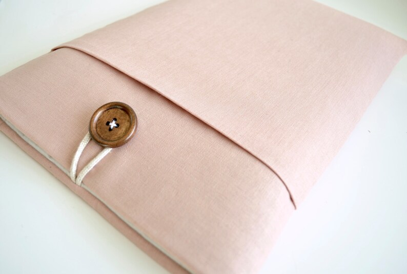 May include: A light pink fabric laptop sleeve with a wooden button closure and a white cord.
