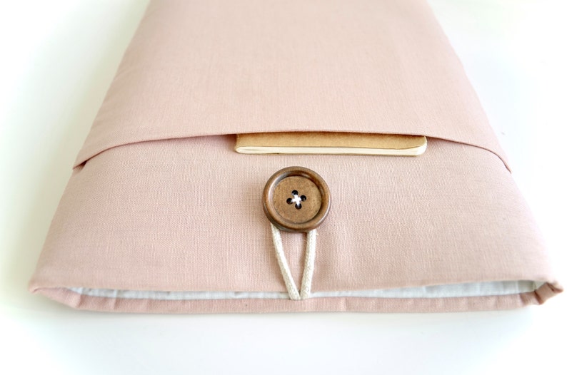May include: A blush pink fabric laptop sleeve with a wooden button closure and a brown notebook peeking out from the top.