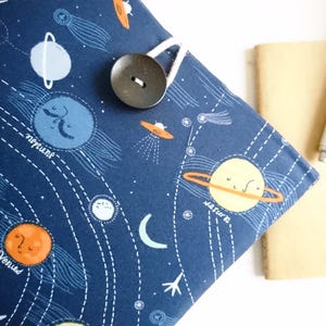 Puede incluir: Una cubierta de libro azul marino con un diseño del sistema solar, con planetas y cometas. Un botón grande y oscuro asegura la cubierta. Un pequeño cuaderno beige y un bolígrafo blanco están al lado.