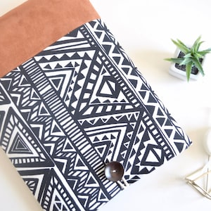 May include: A black and white geometric patterned laptop sleeve with a brown suede flap and a wooden button closure.