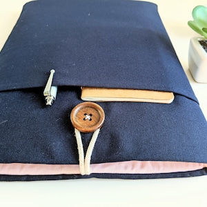 May include: A navy blue laptop sleeve with a brown button closure and a pocket for a pen and notebook. The sleeve has a pink lining.