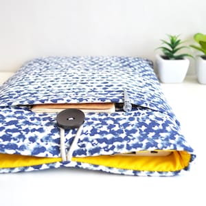 May include: A blue and white patterned laptop sleeve with a yellow lining and a black button closure. The sleeve is closed and has a pen and notebook inside.