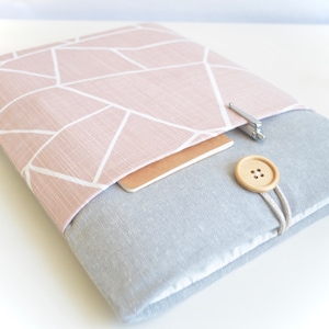 以下が含まれることがあります： 幾何学模様のピンクとグレーのラップトップスリーブ。スリーブはボタン留めのフラップと前面に小さなポケットが付いています。