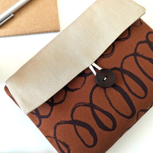 May include: A brown canvas clutch with a beige flap and a wooden button closure. The clutch has a pattern of brown swirls on a brown background.