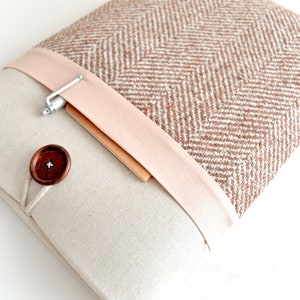 May include: A beige laptop sleeve with a brown and white herringbone pattern on the flap. The flap has a pink fabric band with a silver pen holder and a wooden button closure.