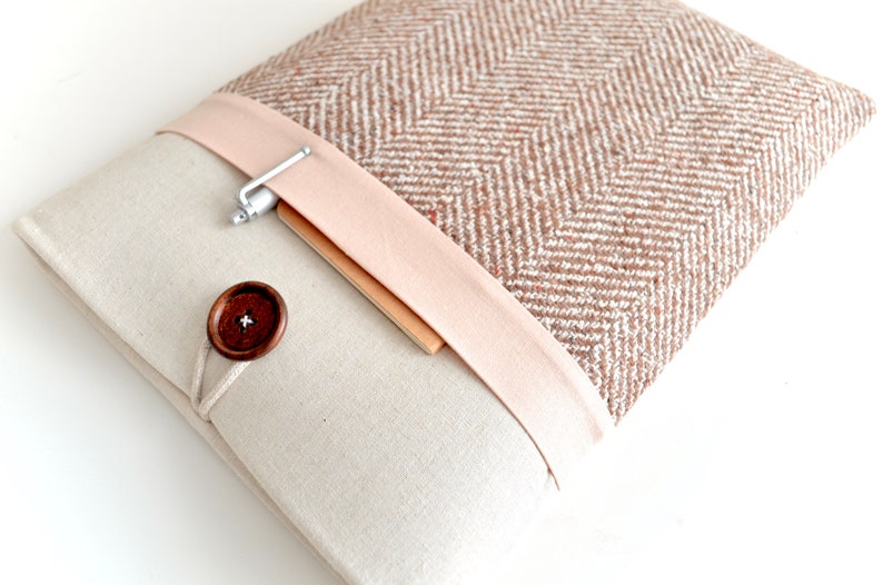 May include: A beige laptop sleeve with a brown and white herringbone pattern on the flap. The flap has a pink fabric band with a silver pen holder and a wooden button closure.