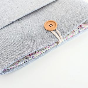 May include: A gray linen laptop sleeve with a floral patterned interior. The sleeve is secured with a wooden button and a cream-colored cord. The interior fabric features a delicate pattern of small flowers.