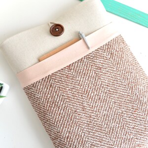 May include: A brown and white herringbone patterned laptop sleeve with a beige linen flap and a wooden button. The flap has a pink fabric trim and a silver pen is tucked into the pocket.