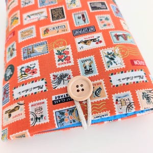 May include: An orange fabric book cover featuring a repeating pattern of colorful postage stamps. The cover has a blue lining and a wooden button closure with a white string. The stamps include text such as "Paris", "Moscow", and "Kenya".