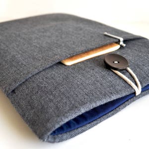 May include: A gray herringbone fabric laptop sleeve with a blue lining and a wooden flap closure. A black button and white string are visible.