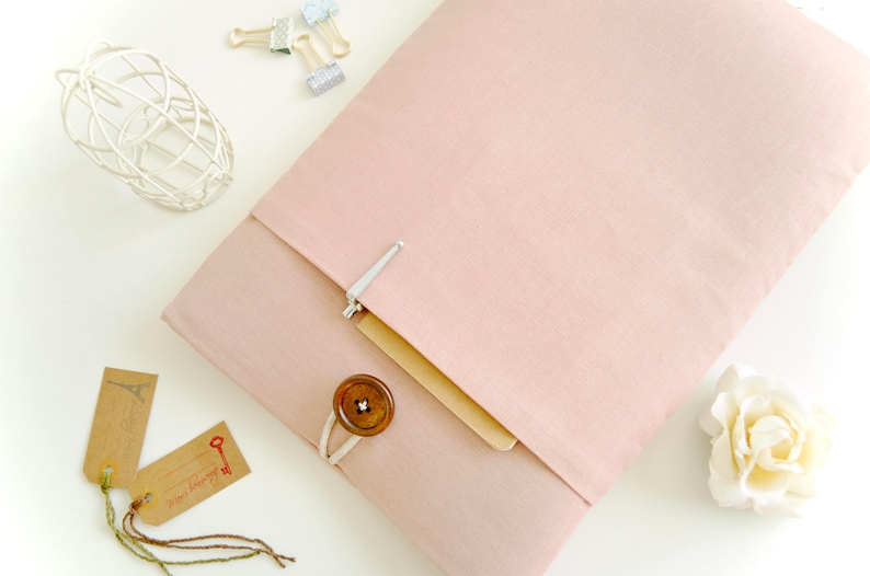 May include: A blush pink fabric laptop sleeve with a flap closure and a button detail. The sleeve is lying on a white surface with a white wire frame and two brown paper tags with a key and Eiffel Tower design.