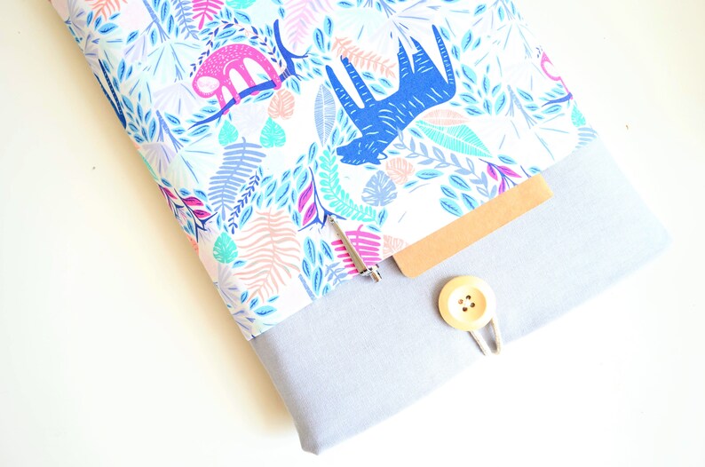 K&ouml;nnte beinhalten: Eine wei&szlig;e Laptoptasche mit einem bunten Dschungel-Druck mit blauen und rosa Tieren, Bl&auml;ttern und Blumen. Die Tasche hat eine graue Klappe mit einem braunen Notizbuch und einem Holzknopf.