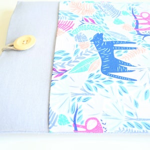 K&ouml;nnte beinhalten: Eine hellgraue Laptoptasche mit einer wei&szlig;en Stoffklappe, die mit einem farbenfrohen Dschungelprint mit blauen Tigern, rosa Affen und gr&uuml;nen Bl&auml;ttern verziert ist. Die Klappe wird mit einem Holzknopf und einer Schnur befestigt.