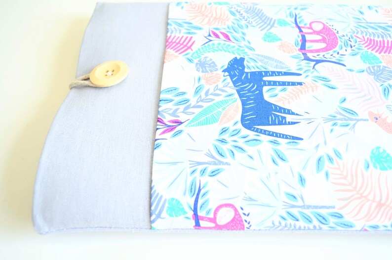 K&ouml;nnte beinhalten: Eine hellgraue Laptoptasche mit einer wei&szlig;en Stoffklappe, die mit einem farbenfrohen Dschungelprint mit blauen Tigern, rosa Affen und gr&uuml;nen Bl&auml;ttern verziert ist. Die Klappe wird mit einem Holzknopf und einer Schnur befestigt.