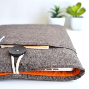 May include: A brown linen laptop sleeve with a white button and a white cord. The sleeve has an orange lining and a pocket on the front. A pen and a notebook are visible in the pocket.
