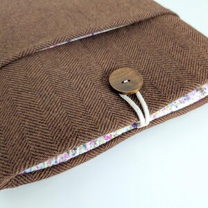 May include: A brown herringbone fabric case with a floral patterned lining. The case is secured with a wooden button and a white cord loop. The case is designed to hold a tablet or other electronic device.
