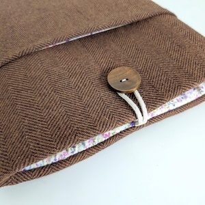 May include: A brown herringbone fabric case with a floral patterned lining. The case is secured with a wooden button and a white cord loop. The case is designed to hold a tablet or other electronic device.