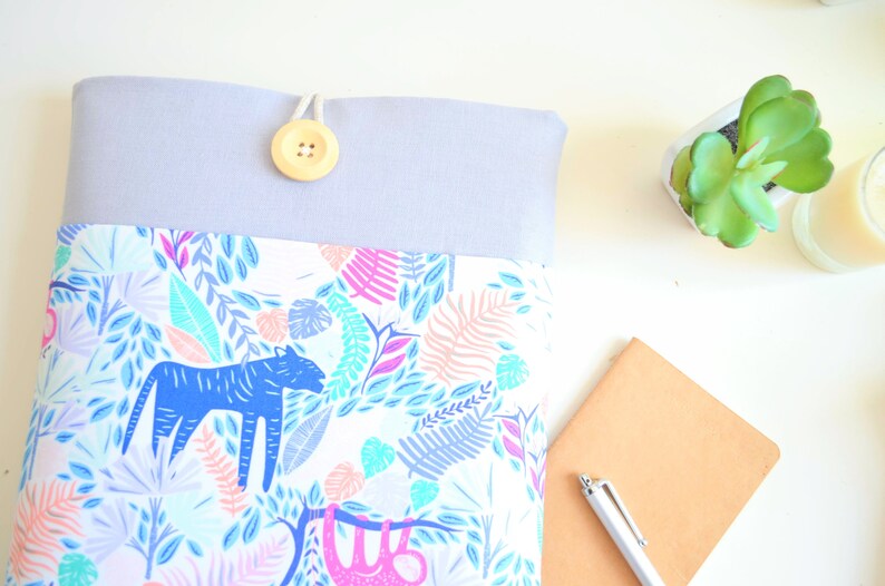 K&ouml;nnte beinhalten: Eine graue und wei&szlig;e Laptoptasche mit einem Dschungel-Druck mit blauen und rosa Tieren und Bl&auml;ttern. Die Tasche hat einen Knopfverschluss und eine hellgraue obere Klappe.