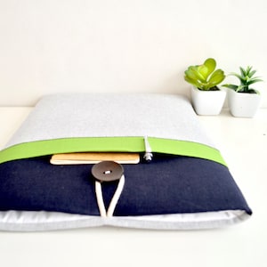 May include: A gray and navy blue laptop sleeve with a green stripe and a brown button closure. The sleeve is lying on a white surface.