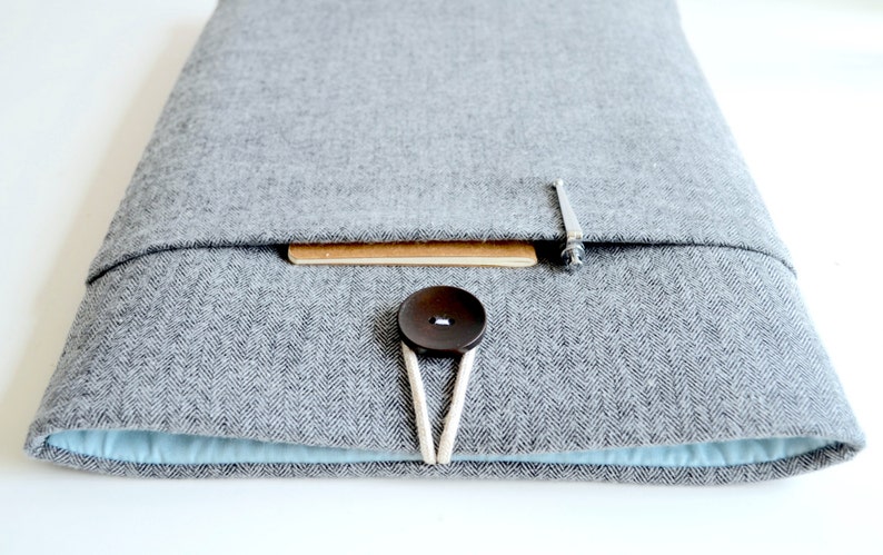 Puede incluir: Una funda gris para port&aacute;til con un patr&oacute;n de espiga, con un cierre de bot&oacute;n marr&oacute;n grande y un interior azul claro. La funda tiene un peque&ntilde;o acento de madera y un cierre met&aacute;lico.