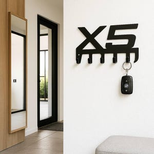 Könnte beinhalten: Schwarzer Metall-Schlüsselhalter in Form von "X5" mit fünf Haken. Ein Autoschlüssel hängt am unteren Haken. Der Schlüsselhalter ist an einer weißen Wand befestigt.
