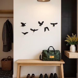 May include: A contemporary hallway scene with a wooden bench and six pairs of shoes. Above, a green duffel bag rests beneath black bird wall art. A black pendant light hangs from the ceiling. The wall is white.