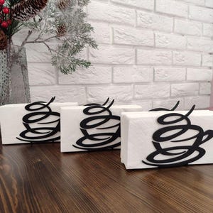 Könnte beinhalten: Drei schwarze Metall-Serviettenhalter in Form von Kaffeetassen, jeder mit einer weißen Serviette. Die Halter stehen auf einem dunklen Holztisch.