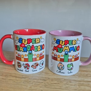May include: Two white ceramic mugs with red and pink interiors. The mugs feature a Super Mario Bros. theme with the text "Super Granddaddie" and "Super Mummi" on each mug. The mugs have a red and pink handle, respectively. The mugs are decorated with characters from the Super Mario Bros. video game series.