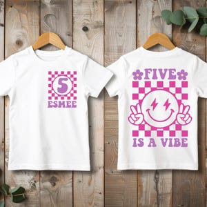 May include: Two white t-shirts with pink and purple graphics. The front features a checkered square with the number 5 and the name Esmee. The back displays a checkered square with a smiley face, peace signs, and the text Five Is A Vibe.