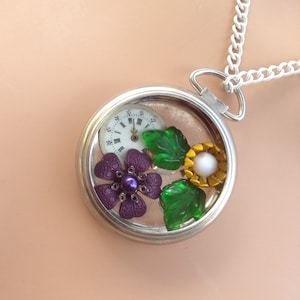 May include: A silver locket necklace with a clock face, purple and green flowers, and a pearl in the center.