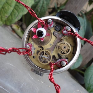 Może przedstawiać: Robot z mechanizmem zegarowym z czerwonymi przewodami i dużym czerwono-czarnym wypukłym okiem. Robot jest wykonany z tarczy zegara z kołami zębatymi i czarną metalową górą.