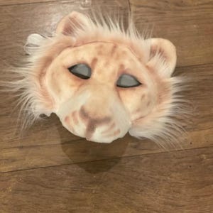 May include: A light brown plush lion mask with white fur around the edges. The mask has black eyes and a pink nose.