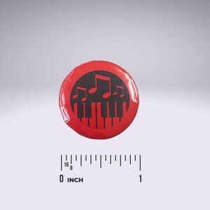 Op de afbeelding: Een rode knop met een zwart ontwerp van een muzieknoot en pianotoetsen. De knop heeft een diameter van 2,5 cm.