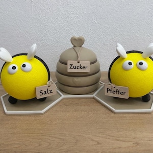 Könnte beinhalten: Ein Set aus zwei gelben bienenförmigen Streuern und einem zentralen beigen bienenstockförmigen Behälter auf einem sechseckigen Sockel. Die linke Biene hat ein Schild mit der Aufschrift Salz, der mittlere Stock Zucker und die rechte Biene Pfeffer.