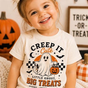 May include: A cream-colored short-sleeve t-shirt featuring a graphic of a ghost holding a pumpkin bucket. The text reads: CREEP IT CUTE, LITTLE GHOST, BIG TREATS, SPOOKY SWEET REPEAT. The design includes a black and white checkered pattern and bats.