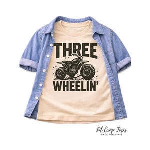 Puede incluir: Una camiseta beige con el texto "THREE WHEELIN'" y un gráfico de un ATV negro. La camiseta se lleva debajo de una chaqueta vaquera azul claro. La camiseta es de Lil Crop Tops.