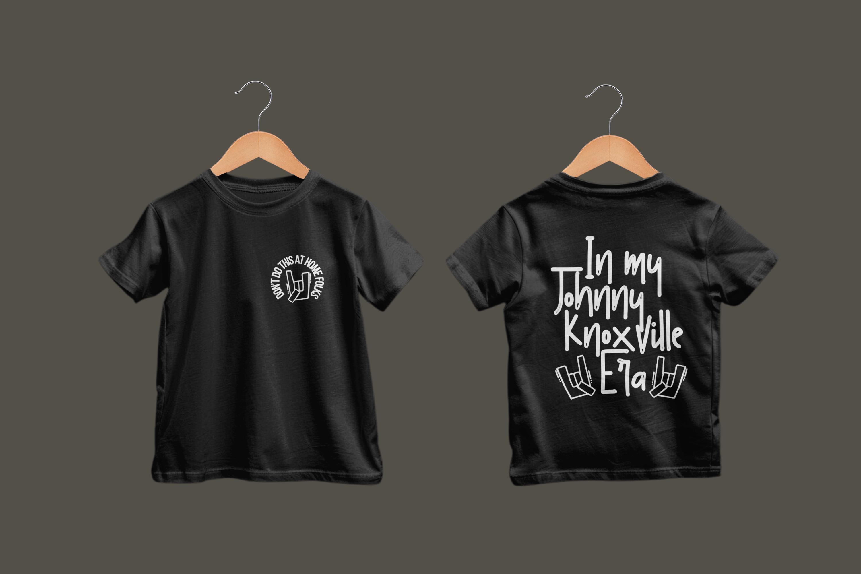 K&ouml;nnte beinhalten: Schwarze T-Shirts auf Holzb&uuml;geln. Auf der Vorderseite des Shirts befindet sich eine wei&szlig;e runde Grafik mit dem Text "DON'T DO THIS AT HOME FOLKS". Auf der R&uuml;ckseite des Shirts steht "In my Johnny Knoxville Era" mit Handgesten.