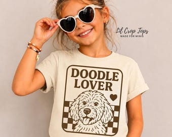 May include: A cream-colored t-shirt featuring a dark brown graphic of a dog face, a checkered pattern, and the text "DOODLE LOVER" with a small heart. The shirt is worn with heart-shaped sunglasses, a beaded bracelet, and a lightning bolt charm.