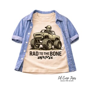 May include: A light tan t-shirt with a graphic of a monster truck driven by a skeleton wearing a baseball cap. The text "RAD TO THE BONE" is below the truck. The shirt is layered under a light blue denim shirt.