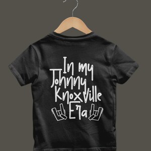 K&ouml;nnte beinhalten: Schwarzes T-Shirt mit dem wei&szlig;en Text "In my Johnny Knoxville Era" und Rock-Handzeichen. Das T-Shirt h&auml;ngt an einem Holzb&uuml;gel.