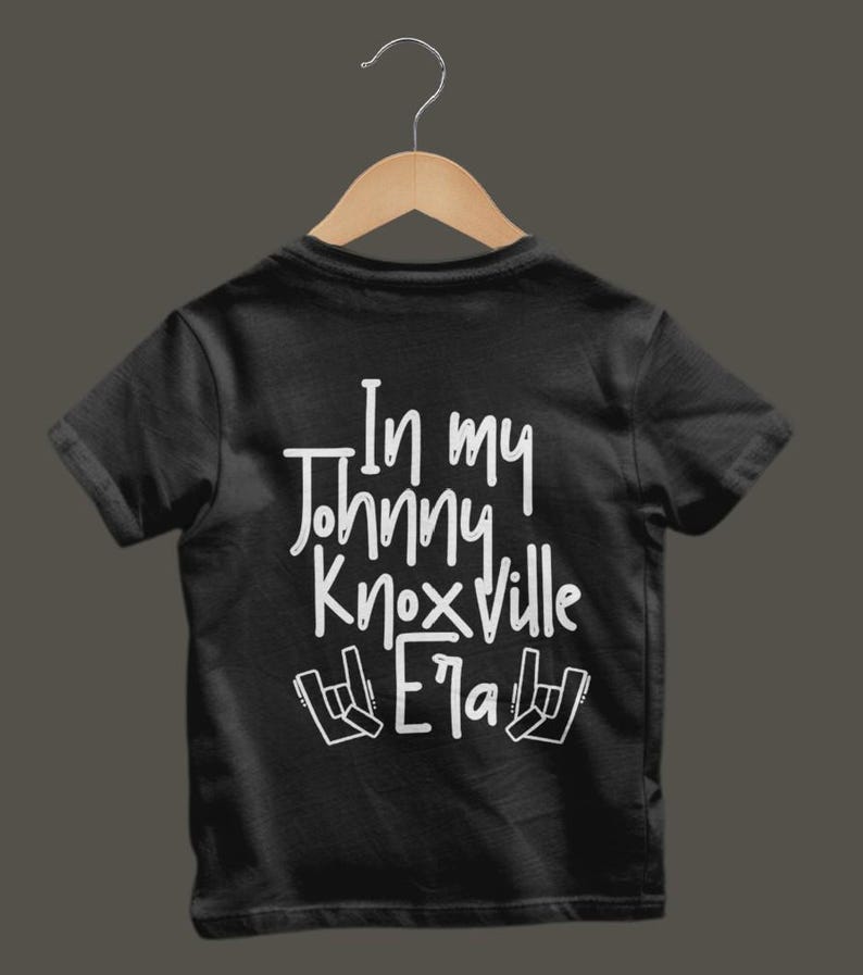 K&ouml;nnte beinhalten: Schwarzes T-Shirt mit dem wei&szlig;en Text "In my Johnny Knoxville Era" und Rock-Handzeichen. Das T-Shirt h&auml;ngt an einem Holzb&uuml;gel.