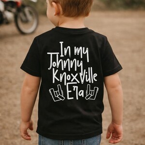 K&ouml;nnte beinhalten: Schwarzes T-Shirt mit dem wei&szlig;en Text "In my Johnny Knoxville Era" und einem Rock'n'Roll-Handzeichen. Das Kind tr&auml;gt eine blaue Jeans. Im Hintergrund ist ein Motorrad zu sehen.