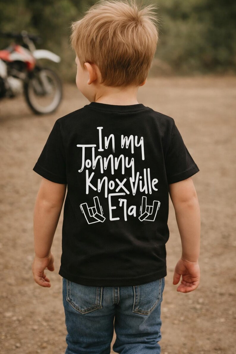 K&ouml;nnte beinhalten: Schwarzes T-Shirt mit dem wei&szlig;en Text "In my Johnny Knoxville Era" und einem Rock'n'Roll-Handzeichen. Das Kind tr&auml;gt eine blaue Jeans. Im Hintergrund ist ein Motorrad zu sehen.