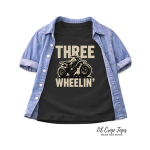 Puede incluir: Camiseta negra con el texto "THREE WHEELIN'" y un gráfico de un ATV de tres ruedas. La camiseta está superpuesta a una camisa vaquera azul claro. El texto "Lil Crop Tops MADE FOR MINIS" está en la parte inferior.