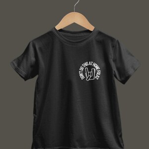 K&ouml;nnte beinhalten: Schwarzes T-Shirt mit wei&szlig;em Aufdruck. Der Aufdruck zeigt den Text "DON'T DO THIS AT HOME FOLKS" in einem Kreis und ein Handzeichen. Das T-Shirt h&auml;ngt an einem Holzb&uuml;gel.