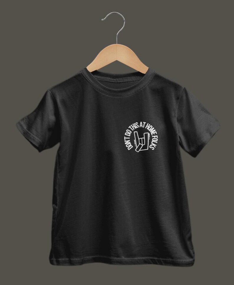 K&ouml;nnte beinhalten: Schwarzes T-Shirt mit wei&szlig;em Aufdruck. Der Aufdruck zeigt den Text "DON'T DO THIS AT HOME FOLKS" in einem Kreis und ein Handzeichen. Das T-Shirt h&auml;ngt an einem Holzb&uuml;gel.