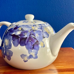 May include: White porcelain teapot with a floral design featuring blue and purple hydrangeas.