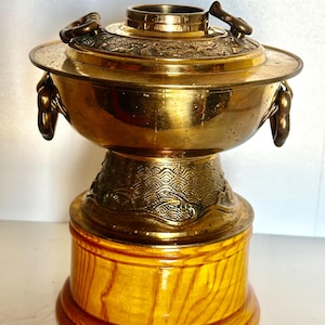 May include: A polished brass incense burner featuring a wide bowl with side ring handles and a lid decorated with intricate relief carvings. The burner sits on a tapered brass base with a wave pattern, mounted on a circular light wood stand.