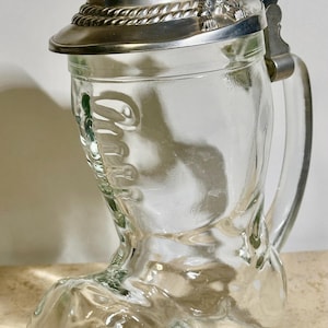 May include: Clear glass beer stein shaped like a boot, with a hinged metal lid and handle. The lid has a decorative rope-like trim and a small brush. The stein is designed to resemble a traditional German boot.